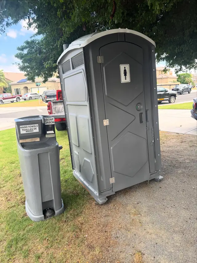 Gray standard porta potty with a freestanding hand-wash station set on grass